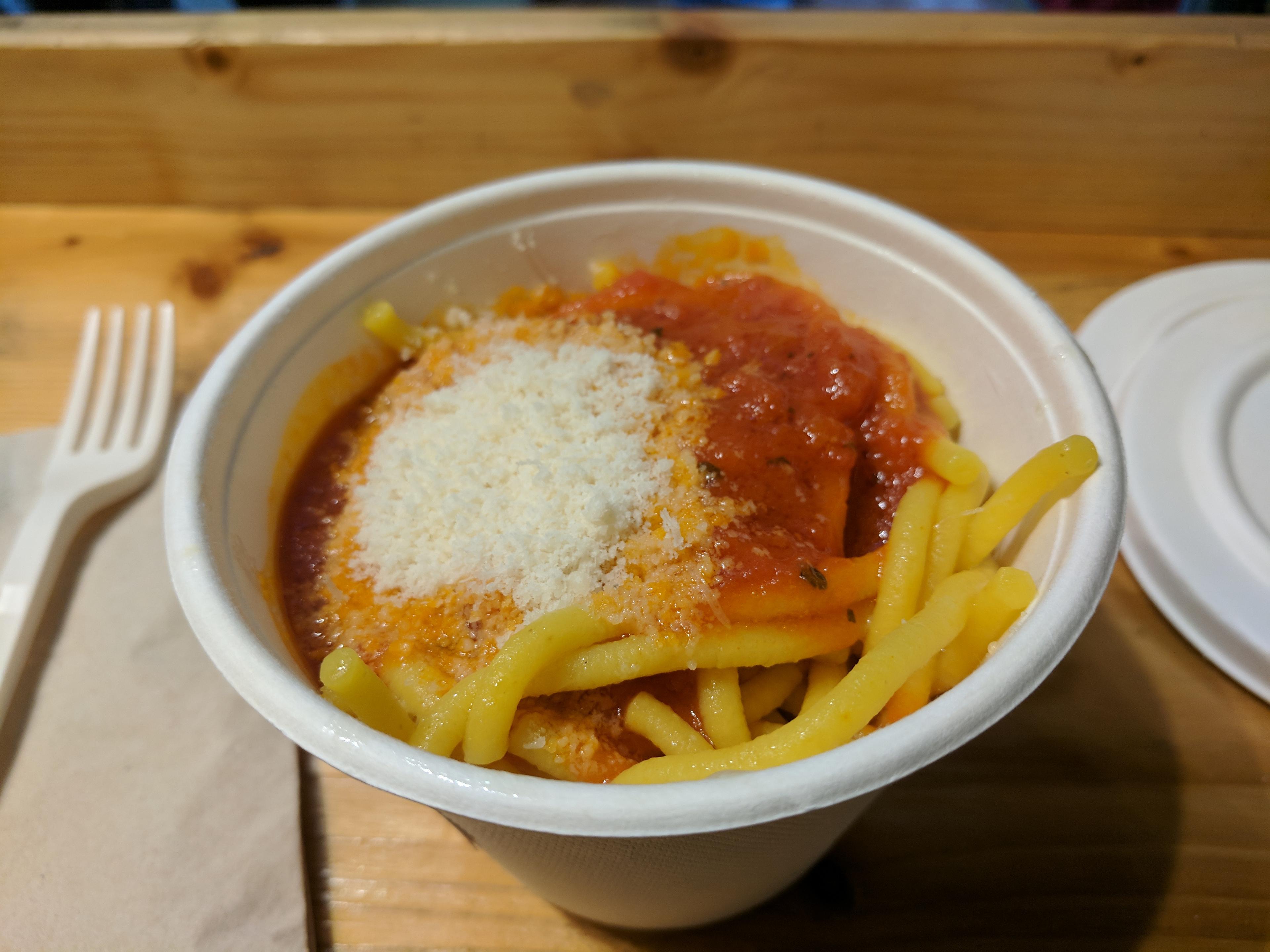 A paper bowl of noodles with red sauce.